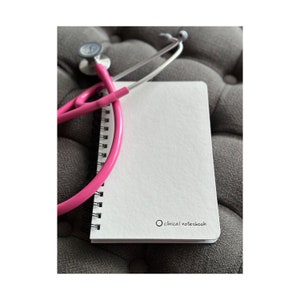 Może przedstawiać: Biały notes w oprawie spiralnej z napisem "clinical notebook" na okładce. R&oacute;żowy stetoskop jest owinięty wok&oacute;ł notesu.