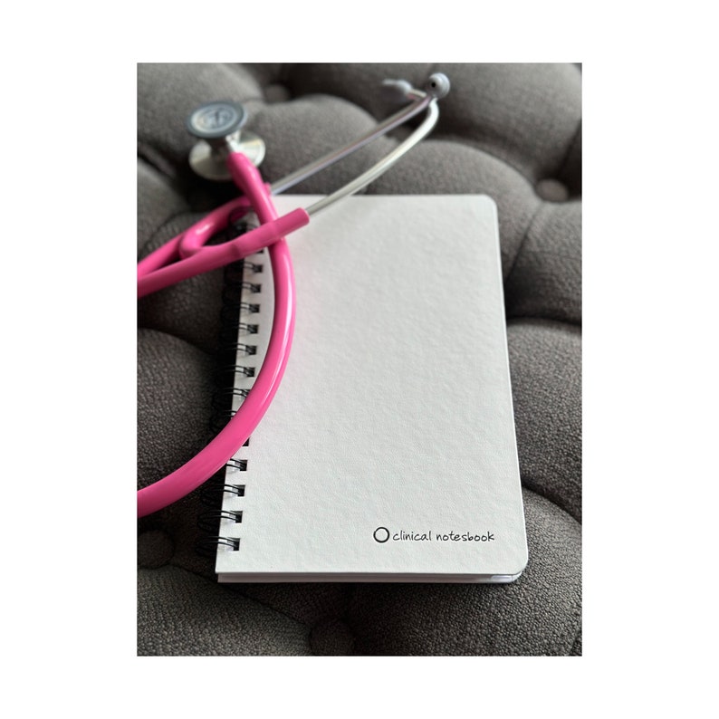 Może przedstawiać: Biały notes w oprawie spiralnej z napisem "clinical notebook" na okładce. R&oacute;żowy stetoskop jest owinięty wok&oacute;ł notesu.