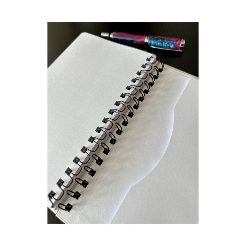 Może przedstawiać: Biały notes w spirali z czarnym spiralnym oprawieniem. Notes jest otwarty na pustej stronie. Czarny i r&oacute;żowy długopis leży na notesie.