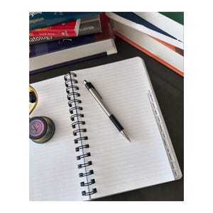 Może przedstawiać: Biały notes w spirali z liniowanymi stronami i srebrnym długopisem leżącym na g&oacute;rze. Notes ma zakładki z boku z etykietami dla r&oacute;żnych przedmiot&oacute;w. Notes leży na ciemnej powierzchni ze stosem książek w tle.