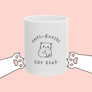 Könnte beinhalten: Ein weißer Keramikbecher mit einer zentralen Illustration eines kleinen weißen Kätzchens mit rosa Wangen. Über dem Kätzchen steht der Text Anti-Social und darunter der Text Cat Club. Zwei weiße Katzenpfoten mit rosa Ballen sind zu sehen.