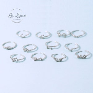 May include: Silver adjustable rings featuring different zodiac constellation designs, each embellished with tiny, sparkling crystals. The rings are presented on a light blue surface, with the "La Lune Accessory" logo visible.