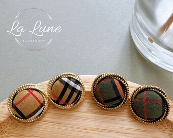 May include: Three round, gold-rimmed buttons with different plaid patterns. The buttons are on a wooden surface.