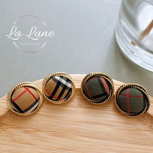 May include: Three round, gold-rimmed buttons with different plaid patterns. The buttons are on a wooden surface.