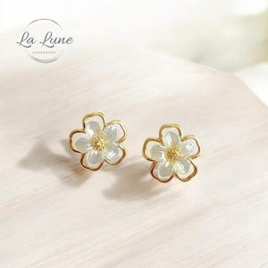 Könnte beinhalten: Ein Paar goldene Blumenohrringe mit weißen, irisierenden Zentren. Die Ohrringe sind klein und zart und perfekt, um jedem Outfit einen Hauch von Eleganz zu verleihen.