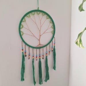 Puede incluir: Un atrapasueños verde con un diseño de árbol de alambre de cobre dentro de un marco circular. El marco está envuelto en hilo verde y adornado con cuentas verdes, cuentas coloridas y borlas verdes.