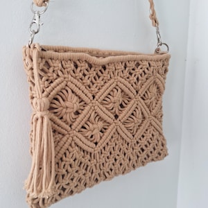 Puede incluir: Bolso de hombro de macramé beige con un patrón geométrico tejido y una borla. El bolso tiene una correa larga con cierres metálicos plateados. El bolso está hecho de un material suave y tejido.
