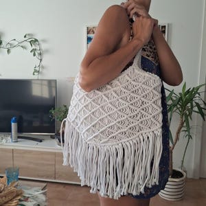 Puede incluir: Un bolso tote de macramé blanco con un patrón geométrico de diamantes y largas borlas. El bolso tiene forma rectangular con un asa. El bolso está hecho de un material tejido blanco roto.
