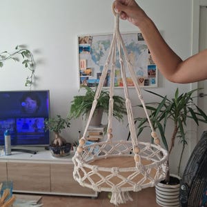 Puede incluir: Colgador de plantas de macramé color crema con base redonda de madera. El soporte para plantas está suspendido de un lazo y presenta un diseño de red tejida con cuentas de madera. La base es de color marrón claro. El colgador de plantas de macramé está diseñado para sostener una planta en maceta.