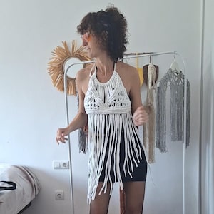 May include: White macrame halter top with long fringe detail. The top is handmade and features a geometric design at the neckline. The top is paired with a black mini skirt. A clothing rack with other macrame pieces is in the background.