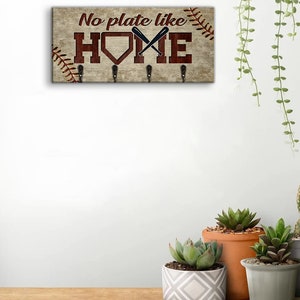 May include: Wooden key holder with a baseball theme. It features a brown background with white stitching, a red home plate, and two crossed baseball bats. The text "No plate like home" is written in red letters.