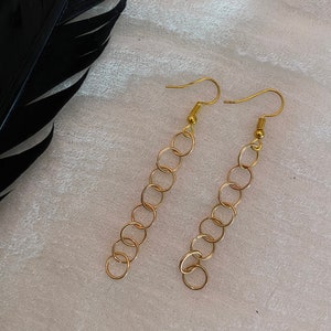 Puede incluir: Un par de pendientes de cadena dorada con un diseño simple de círculo. Los pendientes cuelgan sobre un fondo blanco.
