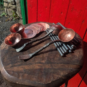 Può includere: Set di utensili da cucina in rame forgiato a mano, tra cui due ciotole, due mestoli con manici attorcigliati e diversi dischi di rame. Gli articoli sono esposti su una superficie di legno scuro, con uno sfondo di legno rosso.