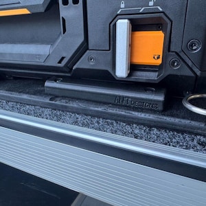 Puede incluir: Una caja de herramientas negra y naranja con un asa plateada y un soporte de metal negro con el texto "ANT Designs" en él. La caja de herramientas está sentada sobre una superficie gris con un borde negro.