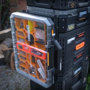 Puede incluir: Una caja de almacenamiento de plástico transparente con múltiples compartimentos, etiquetada como "ToughBuilt Stack-It". La caja está unida a un organizador de herramientas negro y naranja con la etiqueta "ToughBuilt Stack-It".