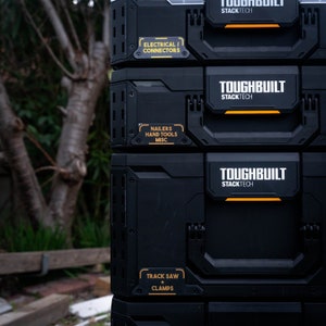 May include: A stack of black toolboxes with yellow labels. The labels read "Toughbuilt Stacktech" and list the contents of each box: "Electrical/Connectors", "Nailers Hand Tools Misc", and "Track Saw + Clamps".