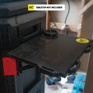 Puede incluir: Una superficie de trabajo negra con acabado de grano de madera se pliega hacia abajo desde un cofre de herramientas negro. La superficie tiene una pegatina amarilla con el texto "ANT DESIGNS" en ella. El texto "TABLETOP NOT INCLUDED!" está impreso en el cofre de herramientas.