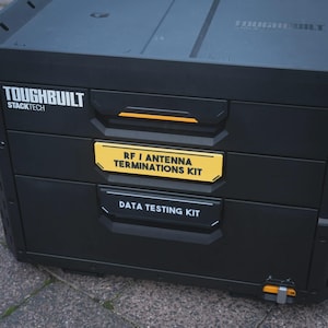 Puede incluir: Caja de almacenamiento de herramientas Toughbuilt Stacktech negra con tres cajones. El cajón superior tiene el texto "RF / ANTENNA TERMINATIONS KIT" en una etiqueta amarilla. El cajón inferior tiene el texto "DATA TESTING KIT". La caja tiene detalles en naranja.