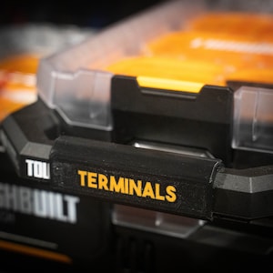 Puede incluir: Una caja de herramientas de plástico negro con una etiqueta amarilla que dice "TERMINALS". La caja de herramientas tiene una tapa de plástico transparente y un asa.