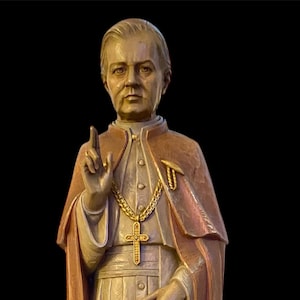 Puede incluir: Estatua de madera de una figura religiosa, con una larga túnica, capa y collar con cruz dorada. La figura sostiene un pergamino con el texto "Quam Singulari". La estatua está sobre un fondo oscuro.