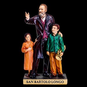 以下が含まれることがあります： 2 人の子供たちとサン・バルトロ・ロンゴの精巧な置物。男性はピンクのシャツの上にダークコートを着ており、片腕を上げています。子供たちはオレンジとグリーンの服を着ています。ベースには「SAN BARTOLO LONGO」と書かれています。