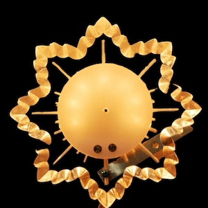 Puede incluir: Un adorno de árbol de Navidad en forma de estrella dorada con un orbe blanco iluminado en el centro. La estrella tiene un tallo de alambre para sujetarlo al árbol.