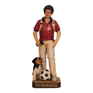 Può includere: Statua di un giovane con polo rosso con disegno di sole, pantaloni color cachi e zaino. È in piedi con le mani in tasca, accanto a un pallone da calcio e a un cane. La base recita "Carlo Acutis."