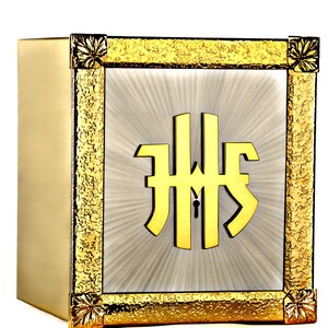 Puede incluir: Una caja de metal dorada con un marco elevado y adornado. La caja tiene un interior blanco con un monograma dorado "IHS" en el centro.
