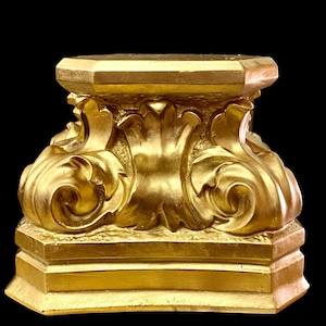 Könnte beinhalten: Ein goldfarbener dekorativer Sockel mit aufwendigen Schnitzereien und einer quadratischen Oberseite. Der Sockel ist aus einem glatten, glänzenden Material gefertigt und hat ein klassisches Design.