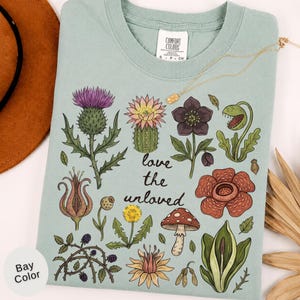 May include: A sage green t-shirt featuring a botanical illustration of various plants, flowers, and a mushroom. The text "love the unloved" is printed in the center. The graphic includes a purple thistle, a cactus, a venus flytrap, and a red mushroom.