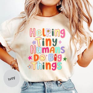May include: A cream-colored t-shirt featuring the multi-colored text Helping Tiny Humans Do Big Things. The design includes various graphic elements such as a smiley face, small flowers, and stars in shades of pink, yellow, blue, and green.