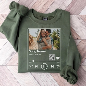 May include: Olive green sweatshirt with a photo of a couple embracing, designed to resemble a music player interface. The interface includes text fields for "Song Name" and "Artist Name", along with play controls and a QR code.