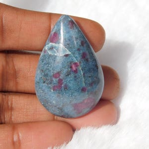 May include: A teardrop-shaped cabochon gemstone with a blue base and red inclusions. The gemstone is a deep blue with a mottled appearance and scattered red spots.
