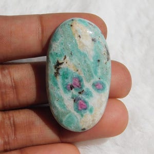 May include: An oval-shaped gemstone with a mix of colors. The stone features a base of light green and white, with small patches of pink and dark spots. The polished surface of the stone is smooth and reflective.