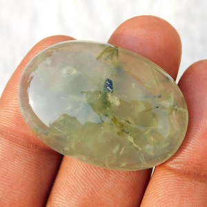Puede incluir: Gema de prehnita translúcida de forma ovalada. La piedra tiene un tono verde pálido con inclusiones verdes más oscuras y negras. La superficie pulida refleja la luz, mostrando la claridad y los patrones internos de la piedra. La gema se sostiene en una mano.