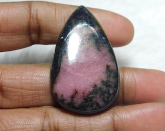 Könnte beinhalten: Ein tropfenförmiger Cabochon aus rosa und schwarzem Rhodochrosit, ein Edelstein, der für seine einzigartigen Farbschattierungen bekannt ist. Der Stein hat eine glatte, polierte Oberfläche und ist in einer silberfarbenen Fassung gefasst.