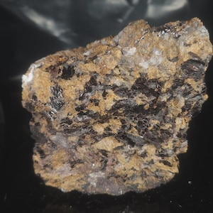 May include: A close-up of a mineral specimen featuring a mix of colors and textures. The rock has a base of tan and brown hues, with patches of black, shiny crystals and small white quartz inclusions. The overall appearance is rough and natural.