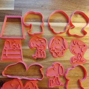 May include: A set of ten pink plastic cookie cutters featuring various Peanuts characters, including Snoopy, Woodstock, and Charlie Brown. The cutters are designed to create different shapes, such as a dog house, a cloud, and a sleeping dog.