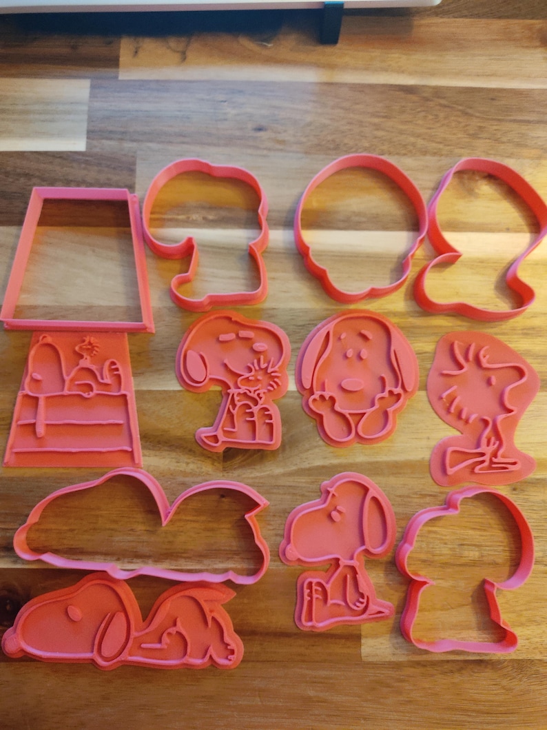 May include: A set of ten pink plastic cookie cutters featuring various Peanuts characters, including Snoopy, Woodstock, and Charlie Brown. The cutters are designed to create different shapes, such as a dog house, a cloud, and a sleeping dog.