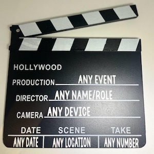 May include: A black and white film clapperboard with the word "HOLLYWOOD" at the top. The clapper has white text fields for "PRODUCTION", "DIRECTOR", "CAMERA", "DATE", "SCENE", and "TAKE". The top clapper section has alternating black and white stripes.