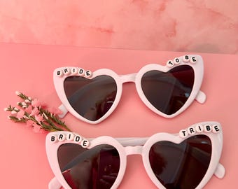 May include: Two pairs of white heart-shaped sunglasses with dark lenses. One pair has the word "BRIDE" spelled out on one side, and "TO BE" on the other. The second pair has "BRIDE" and "TRIBE" on each side. A pink floral accent is on the left.