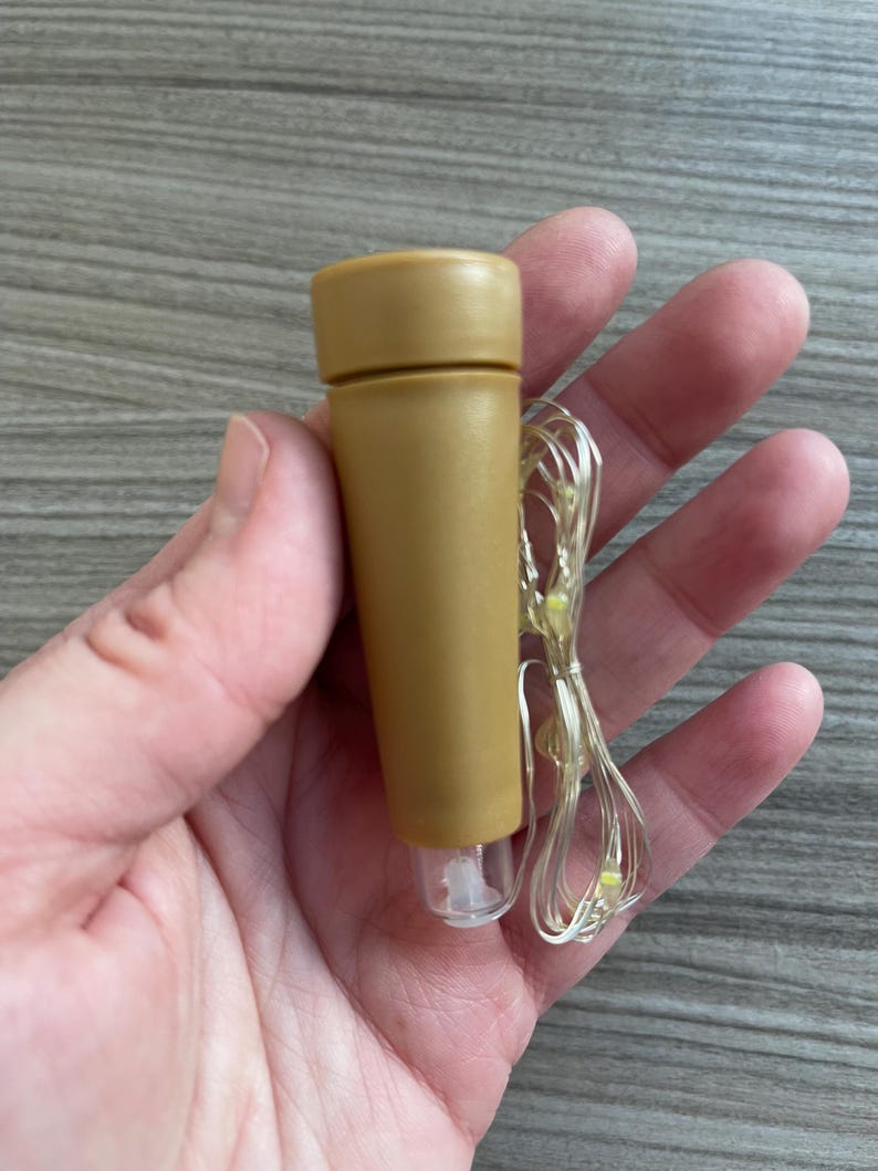 May include: A tan bottle stopper with a cylindrical design, featuring a small clear plastic base and a string of tiny LED lights. The stopper is designed to fit into a bottle opening. The lights are on a thin, flexible silver wire.
