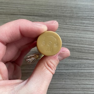 May include: A gold-coloured battery compartment with the text "AAA", "OFF", and "ON". The compartment has a circular design with a directional arrow indicating the on/off switch. The compartment is held in a person's hand.