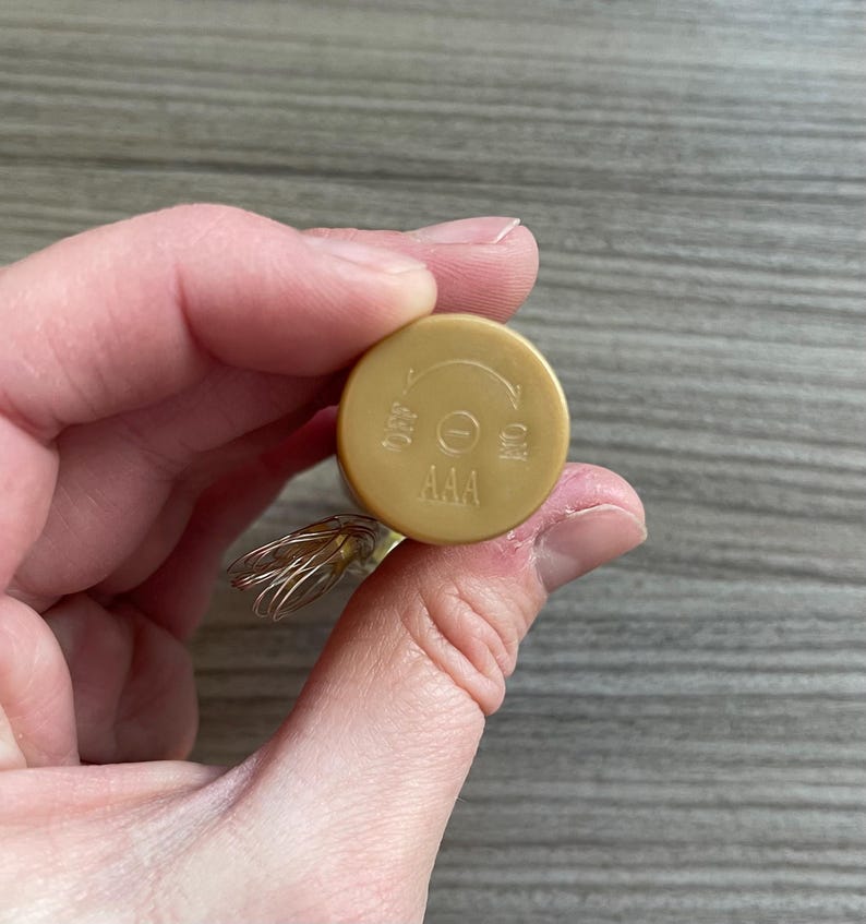 May include: A gold-coloured battery compartment with the text "AAA", "OFF", and "ON". The compartment has a circular design with a directional arrow indicating the on/off switch. The compartment is held in a person's hand.
