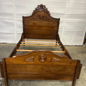 Op de afbeelding: Een donkerbruin houten bedframe met een hoog hoofdeinde en een lager voeteneinde. Beide onderdelen zijn versierd met uitgesneden ronde medaillons en decoratieve accenten. Het frame bevat horizontale houten latten voor ondersteuning.