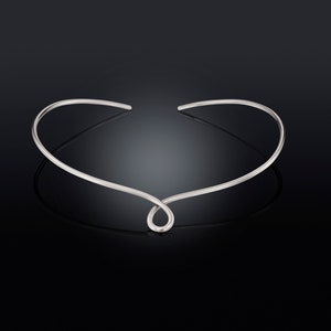 Könnte beinhalten: Eine silberne Choker-Kette mit einem schlichten, eleganten Design. Die Kette besteht aus einem einzigen, durchgehenden Stück Metall, das zu einer Schleife gebogen und dann vorne verdreht wird, um einen einzigartigen und modernen Look zu erzeugen.
