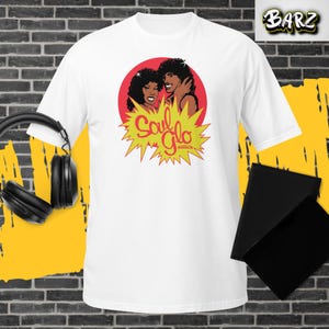 Puede incluir: Camiseta blanca con un gráfico retro de dos personas con cabello rizado dentro de un círculo rojo, con el texto "Soul Glo" en amarillo. El gráfico está rodeado por un diseño de explosión amarilla. La camiseta tiene mangas cortas.