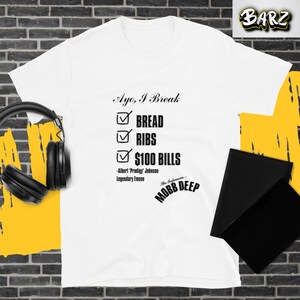 May include: White t-shirt with the text "Ayo, I Break" and a checklist of "BREAD", "RIBS", and "$100 BILLS". The shirt also features the text "-Albert 'Prodigy' Johnson Legendary Emcee" and "MOBB DEEP".