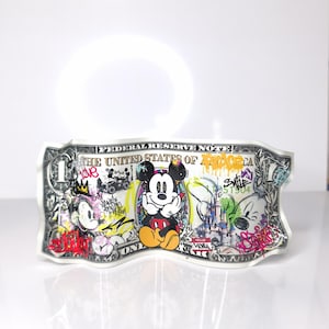 Può includere: Una banconota da un dollaro abbellita con graffiti e personaggi dei cartoni animati, tra cui Topolino. La banconota presenta colori vivaci e aggiunte artistiche, trasformando la valuta in un'opera d'arte unica.