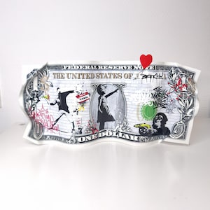 Può includere: Un oggetto d'arte decorativo a forma di banconota da un dollaro ondulata con illustrazioni in stile graffiti in bianco e nero, tra cui un bambino che cerca di afferrare un palloncino a forma di cuore rosso, una scimmia con una banana e la scritta The World Is Mine.
